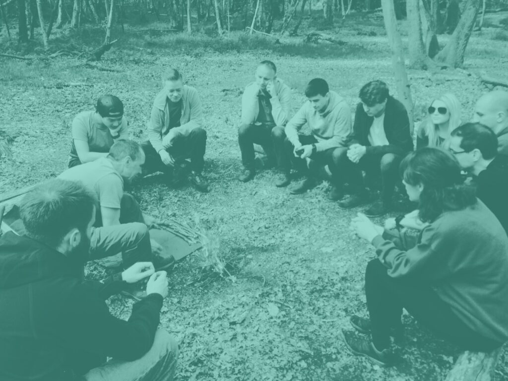 Team sitting in a circle around campfire on bushcraft apprentice challenge day with teal image overlay