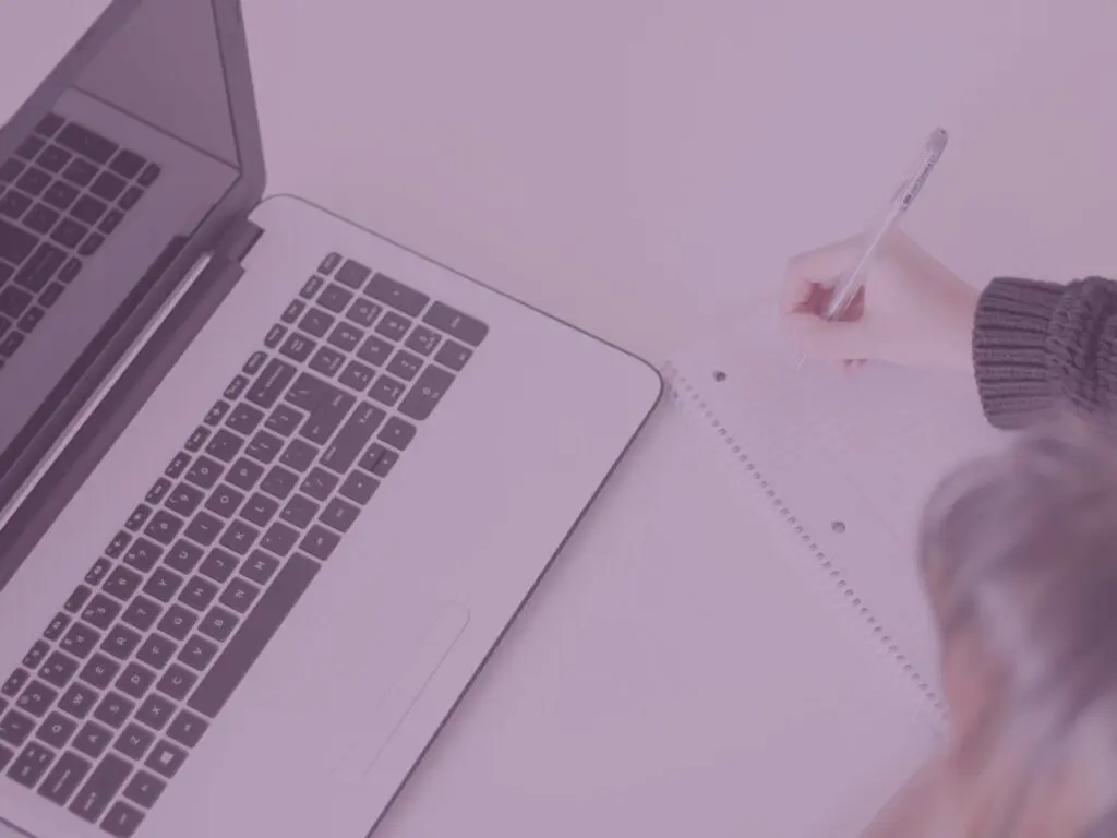 Purple woman writing on notepad next to open laptop