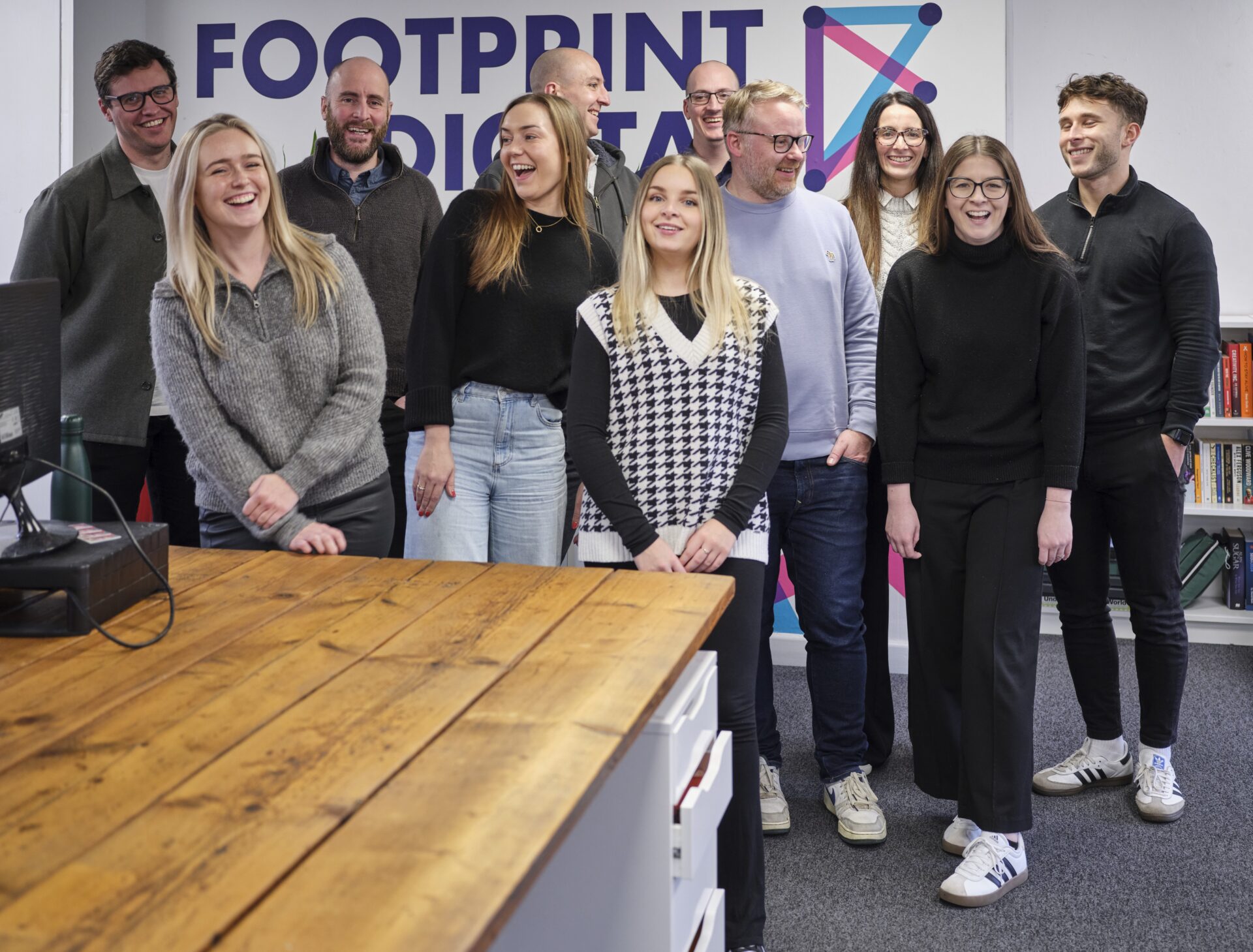 Full colour relaxed and smiling team photo in office stood by Footprint Digital logo